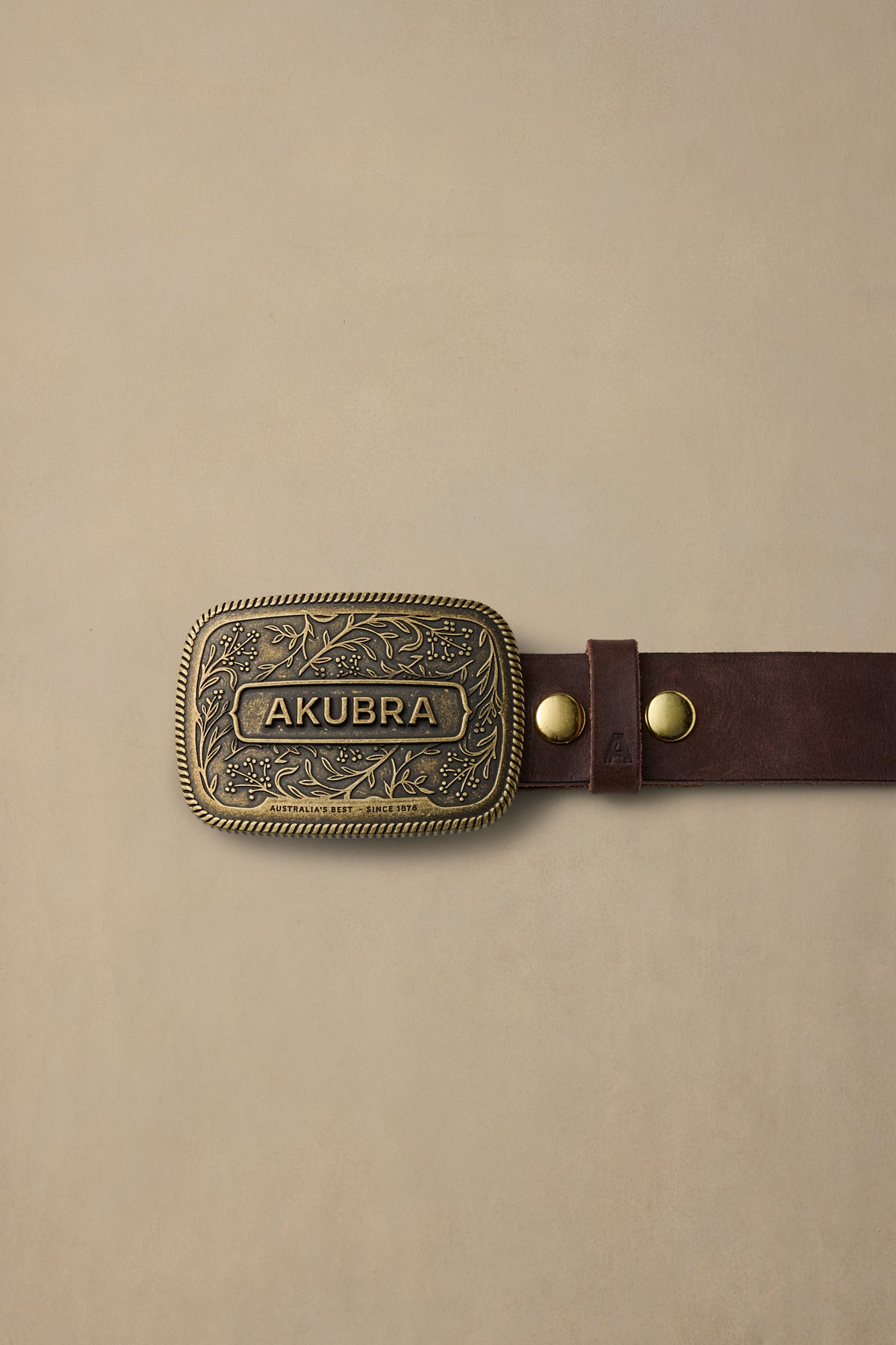 Brass belt buckle with ornate floral design and AKUBRA text on dark brown leather strap against cream background