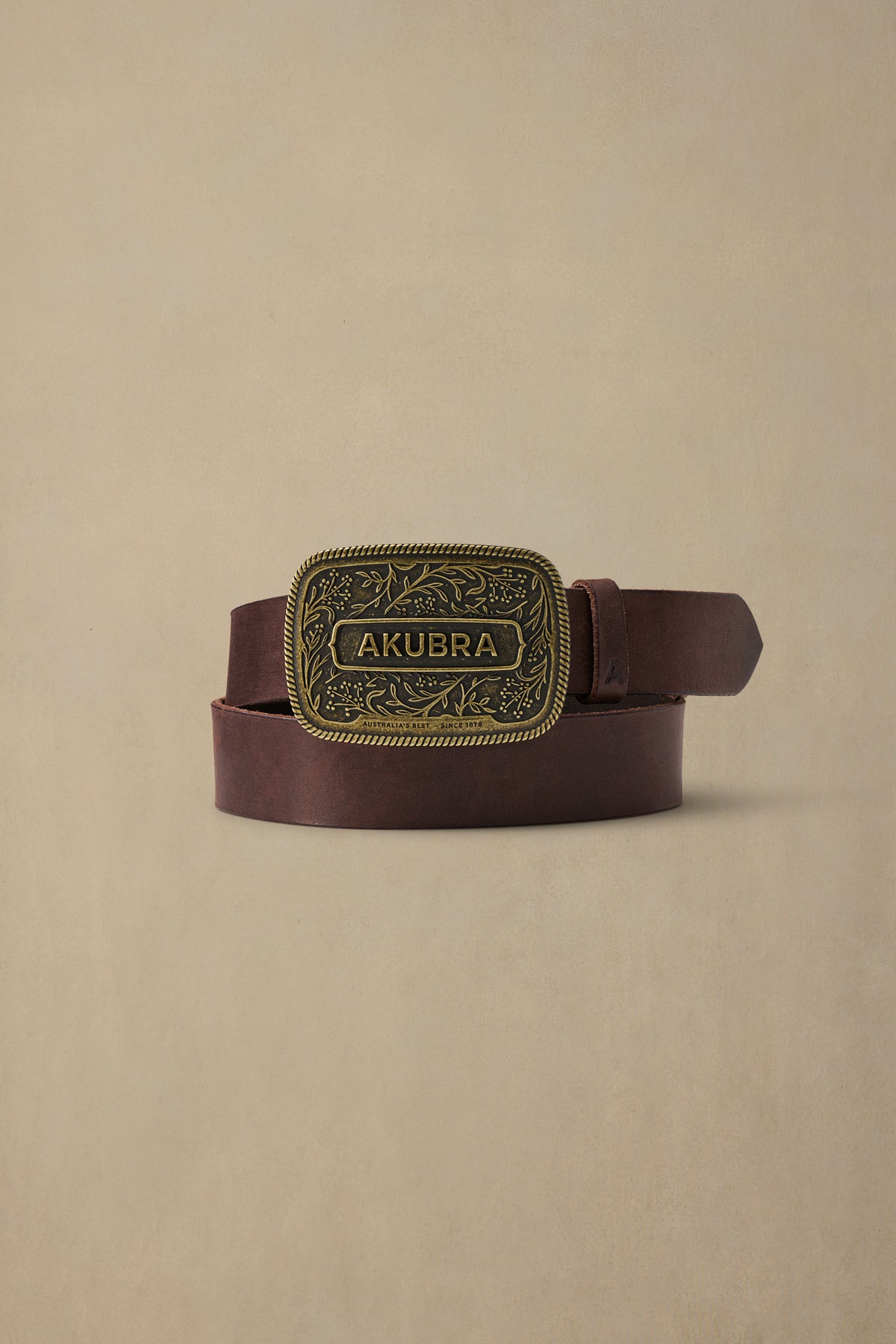 Chocolate brown leather belt with ornate brass buckle engraved with "AKUBRA" text and decorative floral pattern on beige background