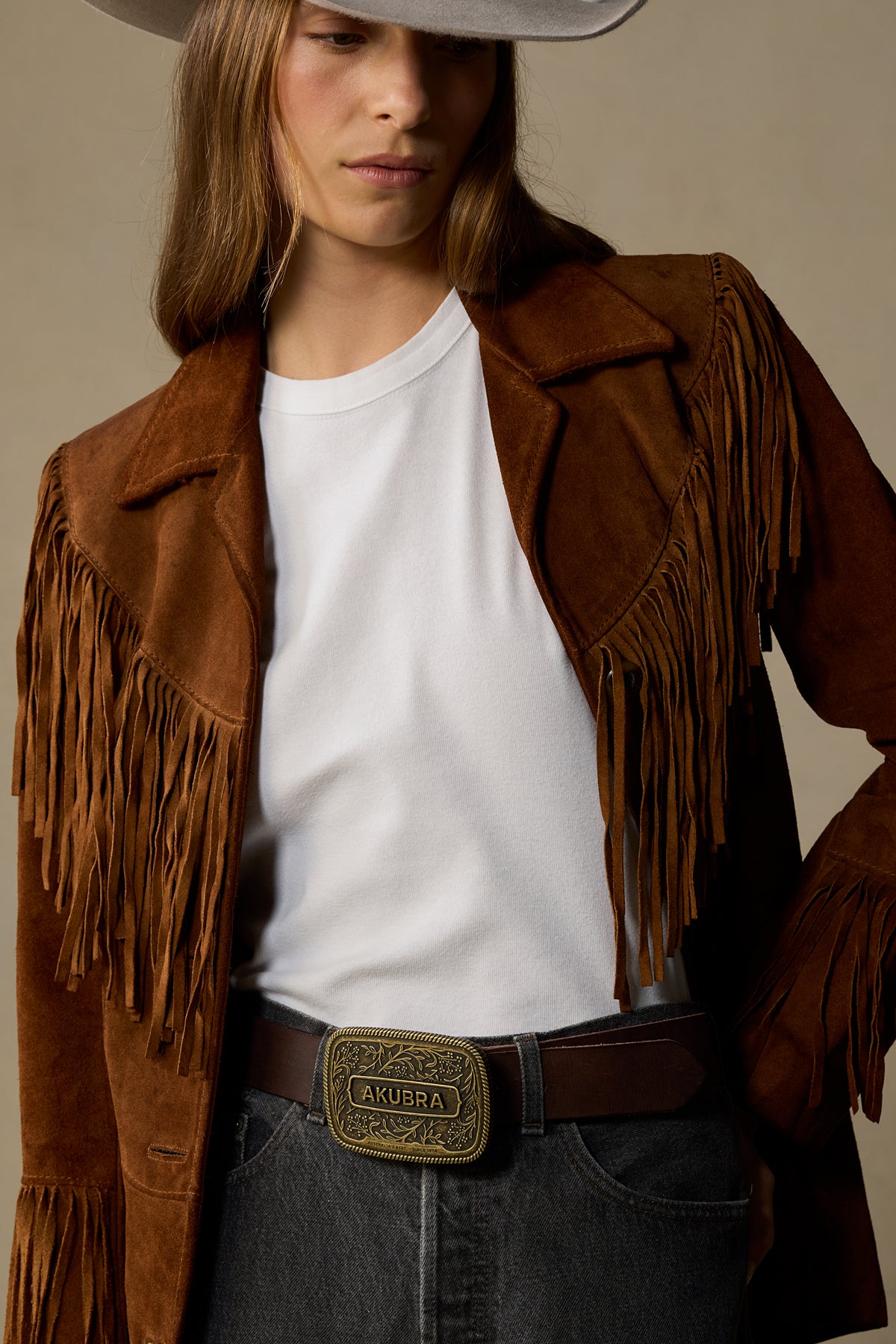 Brown leather fringe jacket with white tee, brass belt buckle, and gray cowboy hat on neutral background