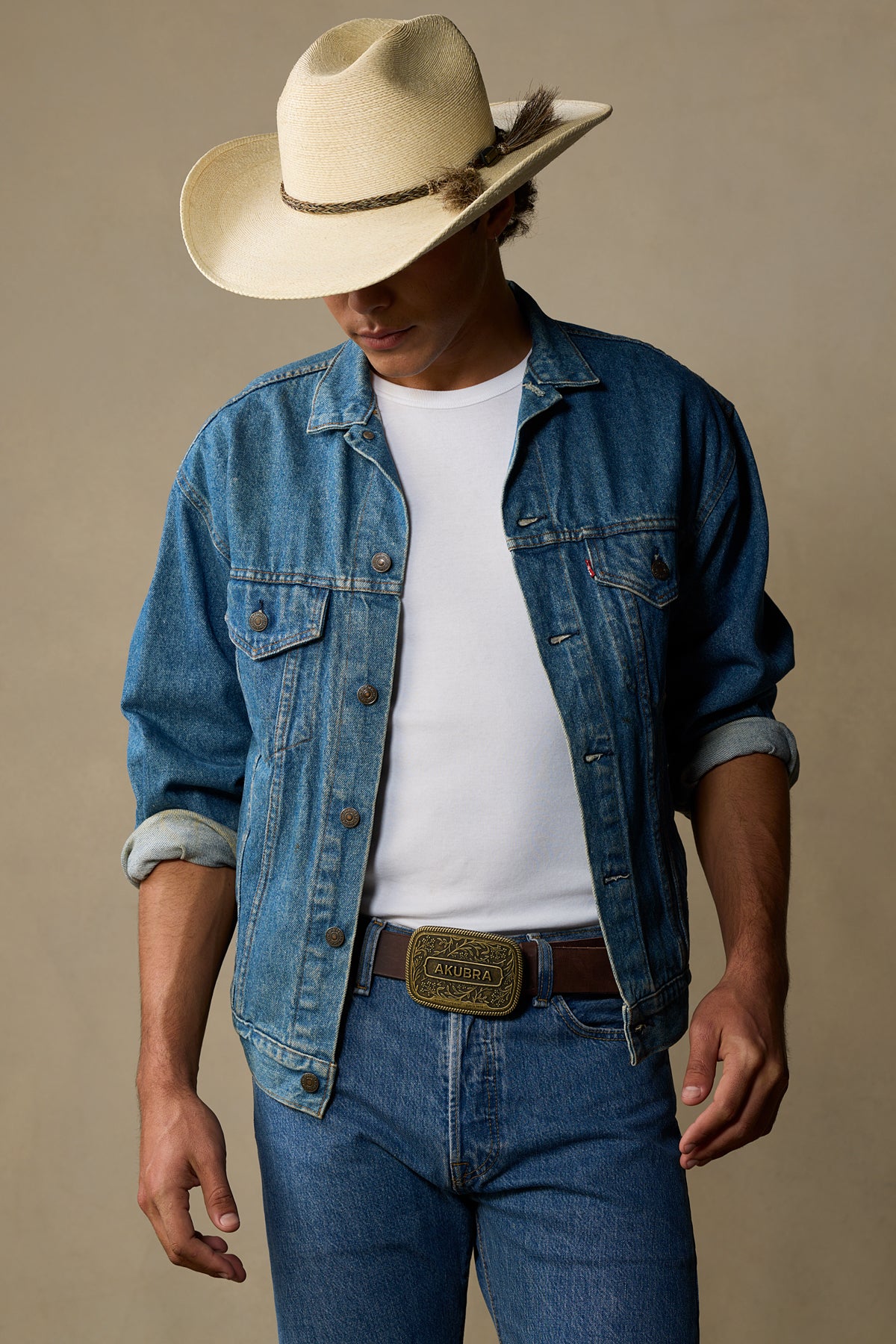 Cream cowboy hat with braided band on model wearing white tee, denim jacket, and brown leather belt with brass buckle