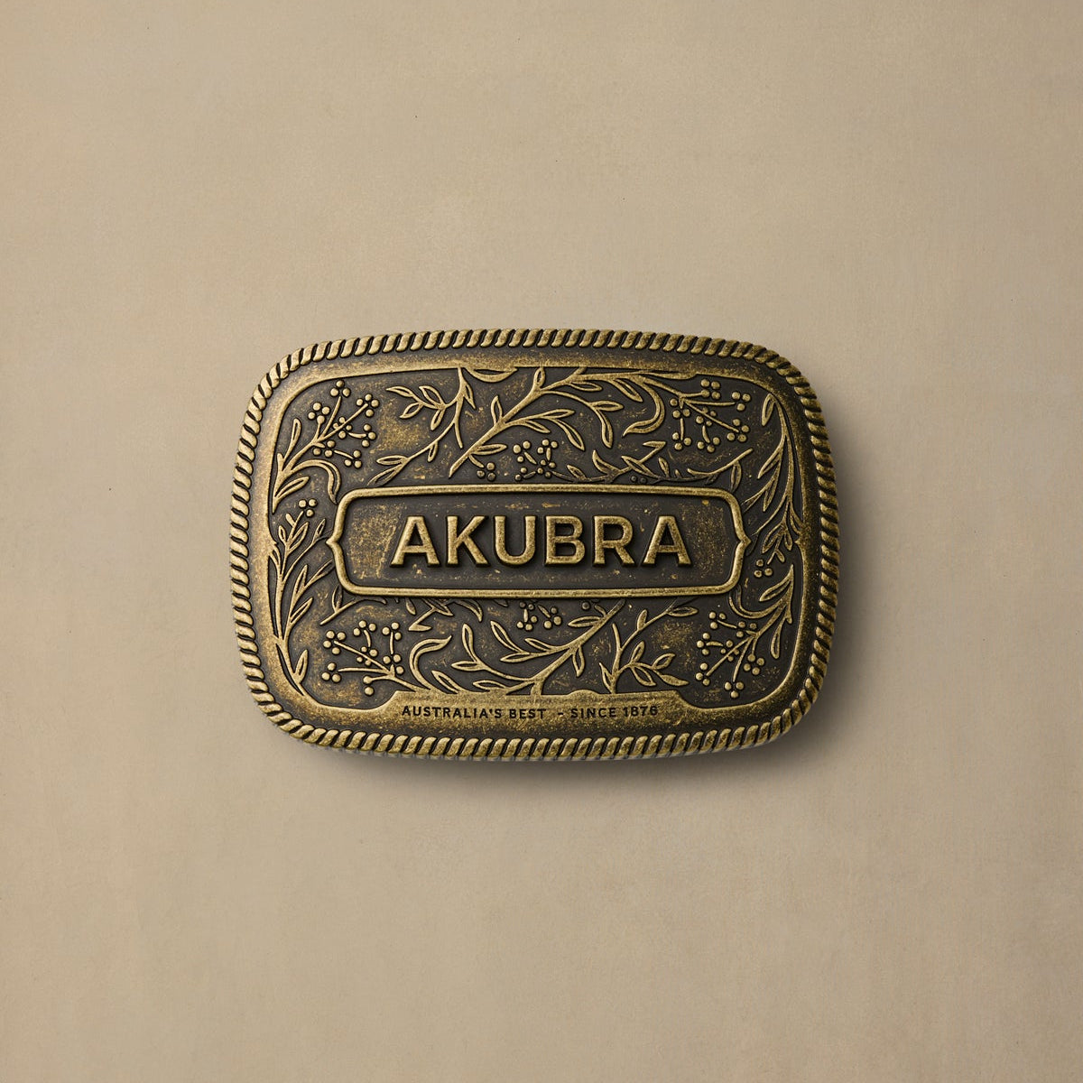 Brass belt buckle with Akubra branding, ornate floral engraving, rope edge border on cream background