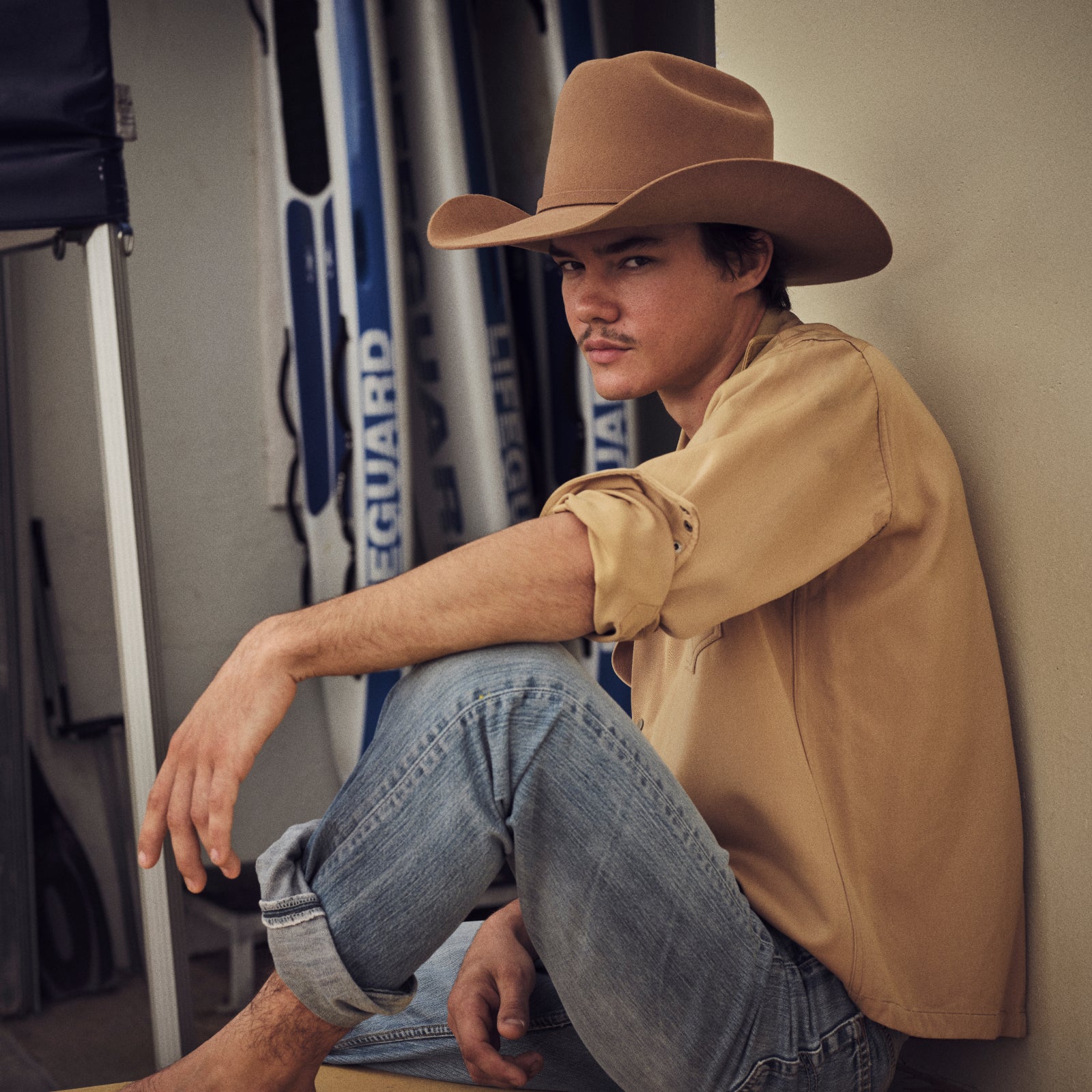 Man wearing a brown cowboy, western Akubra hat and beige shirt sitting against a wall with surf equipment in the background.