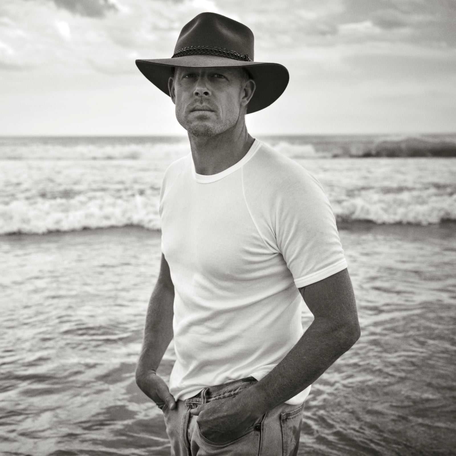 Man (Mick Fanning) wearing a white t-shirt and an Akubra hat standing on a beach with ocean waves in the background.