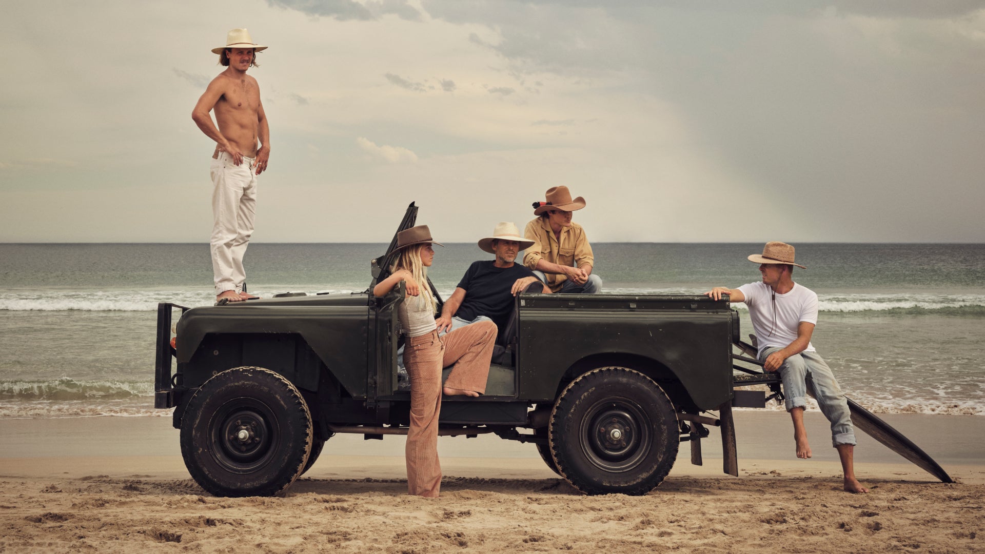 Group of people on a beach with an old-fashioned, vintage land rover truck, wearing Akubra hats
